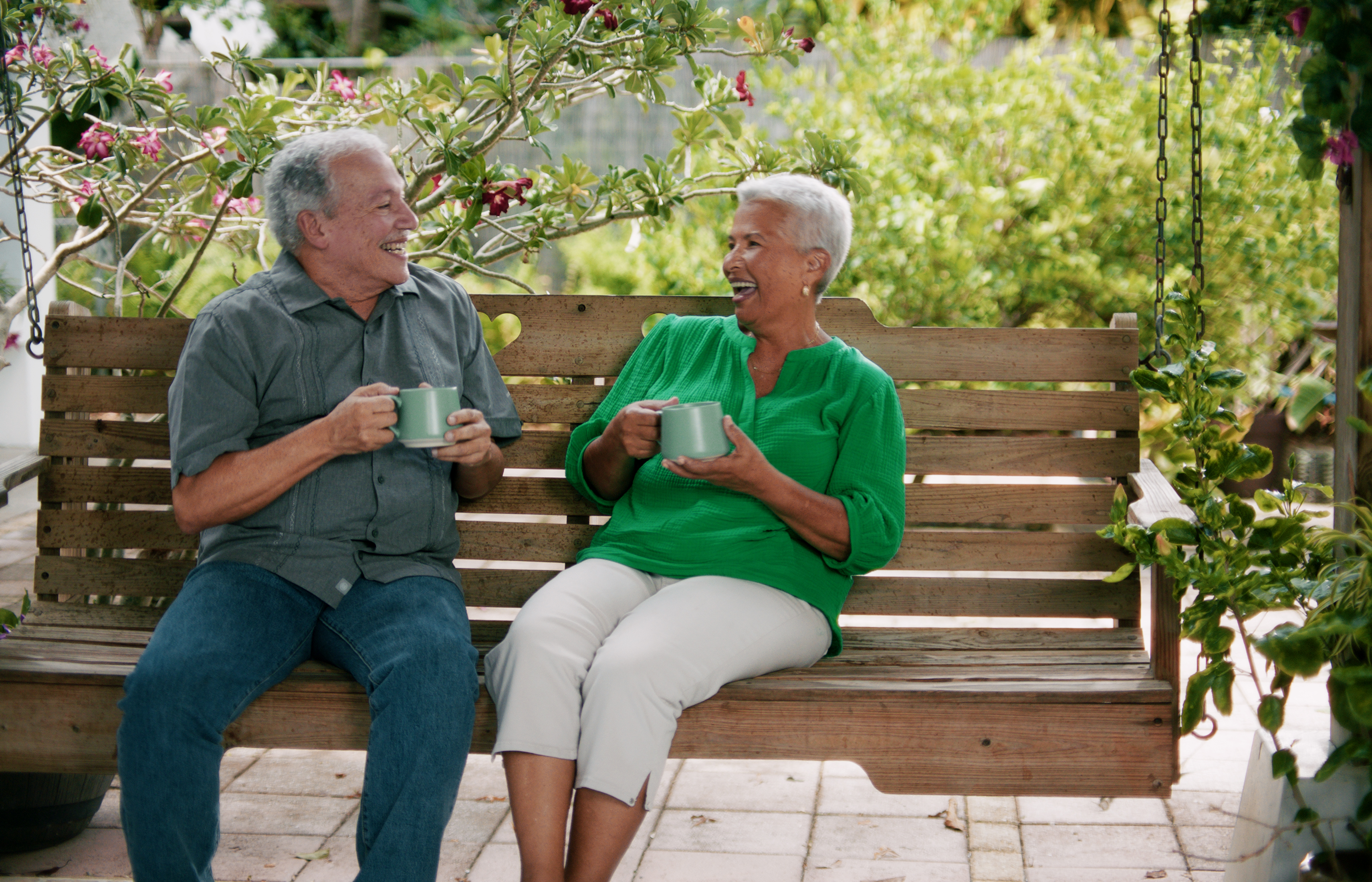 An elderly smiling couple sitting on a wooden garden swing, enjoying a cup of hot beverage while cheerfully talking, surrounded by plants and flowers.